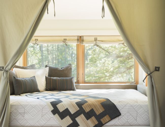 A cozy bed by a window with curtains, pillows, and a patterned blanket; storage shelves are underneath.