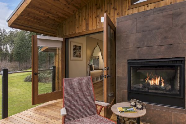 A cozy patio with a wicker chair and small table sits by a modern fireplace, overlooking a green yard, adjoining an inviting interior room.