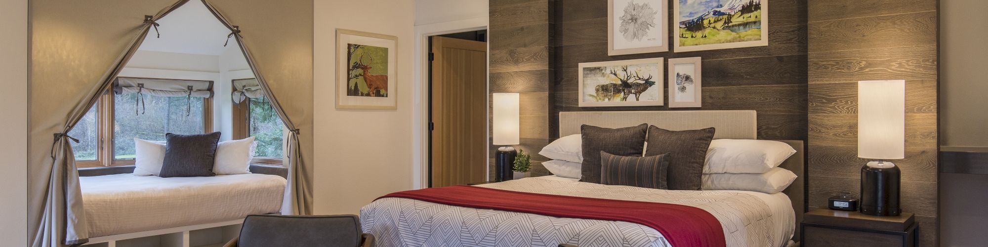 A cozy bedroom with a large bed, two chairs, lamps, and framed art. A window nook features a cushion, enhancing the inviting atmosphere.