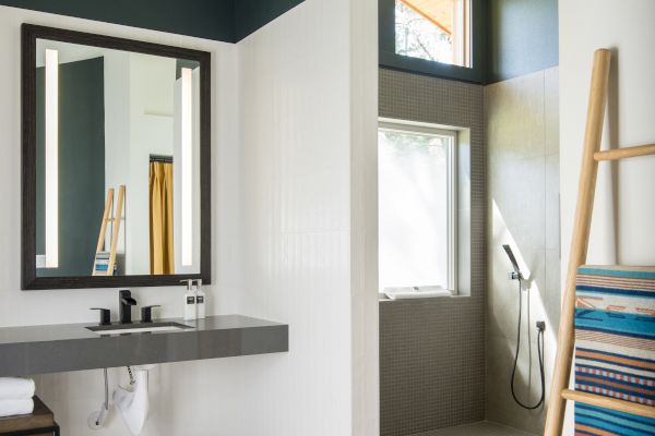 A modern bathroom with a mirror, countertop, walk-in shower, ladder with towels, and two storage baskets underneath.