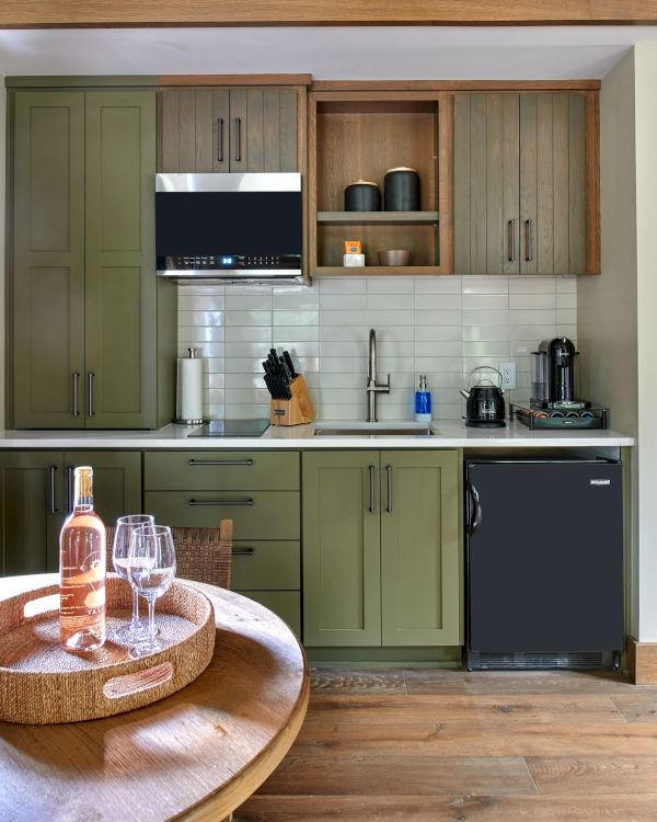 A modern kitchen with olive cabinets, a microwave, sink, espresso machine, and a table with wine and glasses on a tray.