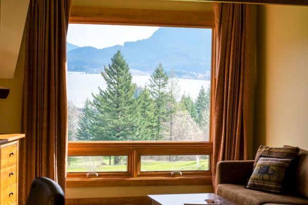 A cozy room with a large window view of trees, mountains, and a lake, featuring a sofa, cushions, and a wooden desk.