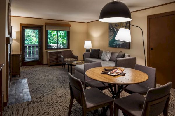 A cozy living room with a round table, chairs, a gray sofa, floor lamp, and window offering a view of greenery.