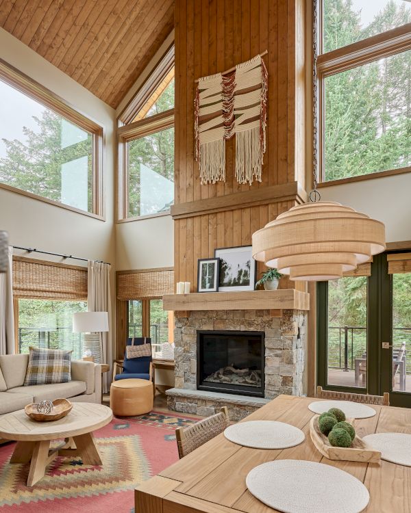 A cozy living room features large windows, a stone fireplace, wood accents, a woven wall hanging, and a stylish dining table.