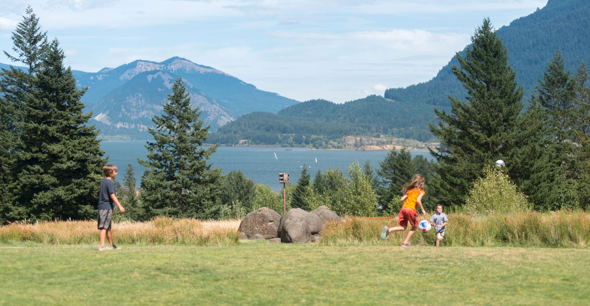 Three kids play soccer on a grassy field with trees and mountains in the background under a partly cloudy sky.
