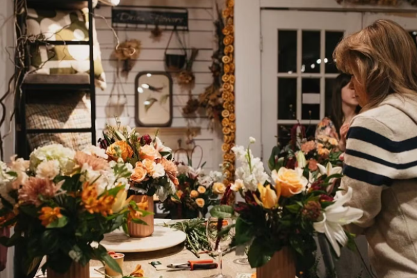 A cozy floral shop scene with two people arranging colorful flower bouquets on a table, surrounded by decor and tools.
