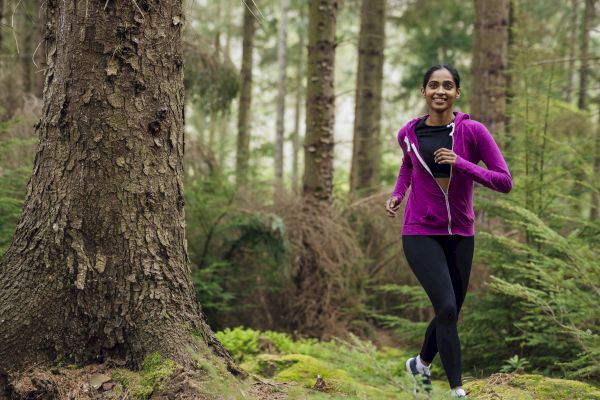 A person jogging in a forest, wearing a purple jacket and black leggings, surrounded by lush green trees and plants.