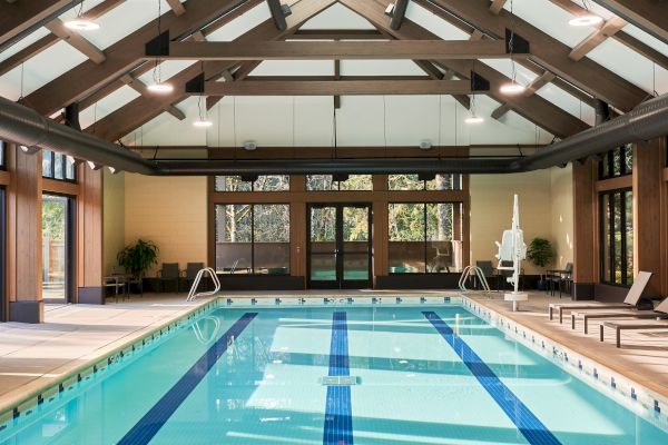 An indoor swimming pool with blue lane markings, wooden beams, skylights, and lounge chairs by the side in a spacious room.