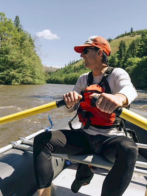 A person is rowing a raft on a river surrounded by trees, wearing a life jacket and cap, under a clear blue sky.