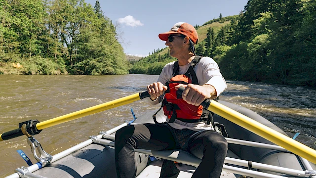 A person is rowing a raft on a river surrounded by trees, wearing a life jacket and cap, under a clear blue sky.