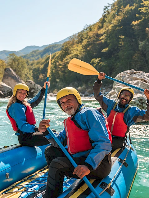 Three people in a raft paddle down a river, wearing helmets and life jackets, surrounded by trees and rocks.