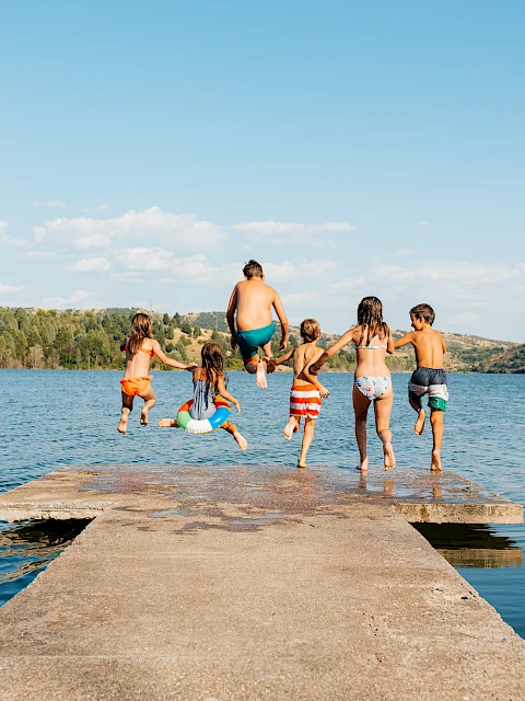 A group of children in swimwear are joyfully jumping off a dock into a lake on a sunny day.