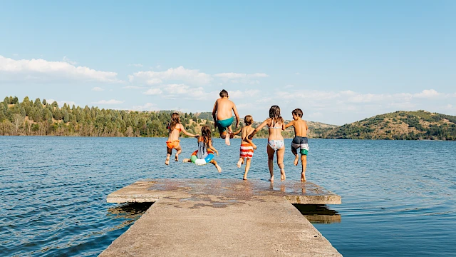 A group of children in swimwear are joyfully jumping off a dock into a lake on a sunny day.