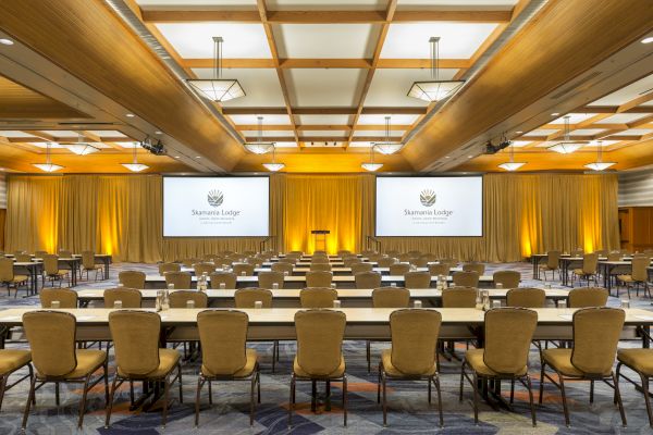 A conference room is set up with rows of tables and chairs facing two large screens displaying a logo.