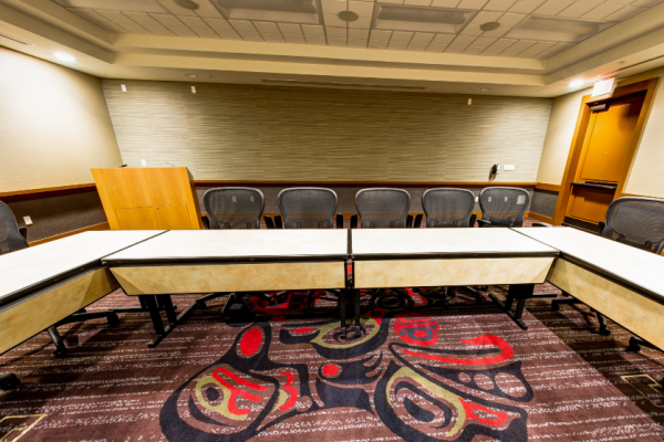 A conference room with a U-shaped table setup, surrounded by chairs, featuring a patterned carpet and neutral walls, ready for meetings.