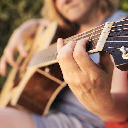 A person is playing an acoustic guitar outdoors, focusing on their hand on the strings.