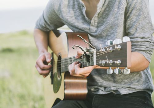 A person playing an acoustic guitar outdoors, wearing a gray long-sleeve shirt with a capo on the guitar neck.