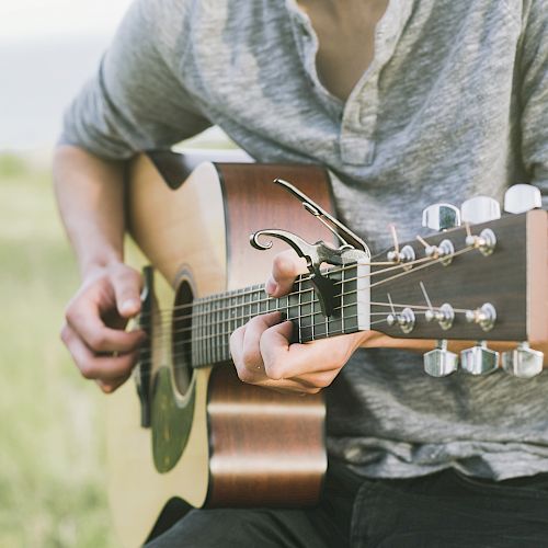 A person playing an acoustic guitar outdoors, wearing a gray long-sleeve shirt with a capo on the guitar neck.