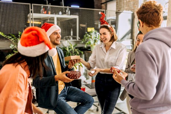 A group of people wearing festive hats are joyfully exchanging gifts in what appears to be an office setting with plants in the background.