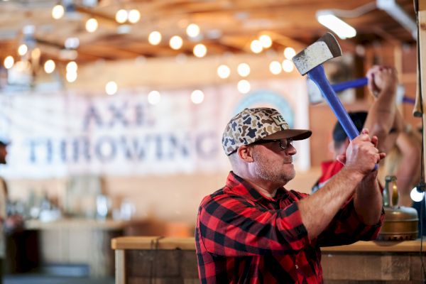 A person in a plaid shirt and cap is preparing to throw an axe in an indoor axe-throwing venue.