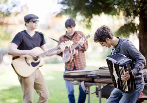 Three people are playing instruments outdoors: a guitar, a mandolin, and an accordion in a park-like setting.