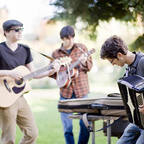 Three people are playing instruments outdoors: a guitar, a mandolin, and an accordion in a park-like setting.