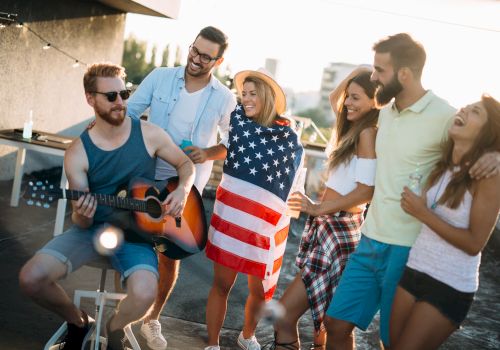 A group of friends is gathered outdoors, enjoying music as one plays guitar, with one person wrapped in a flag, all smiling and having fun.