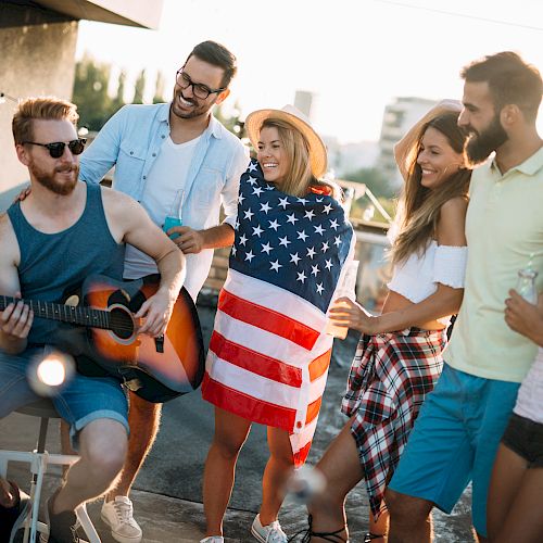 A group of friends is gathered outdoors, enjoying music as one plays guitar, with one person wrapped in a flag, all smiling and having fun.