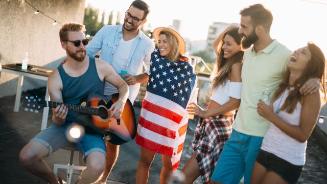A group of friends is gathered outdoors, enjoying music as one plays guitar, with one person wrapped in a flag, all smiling and having fun.