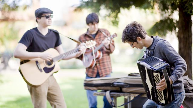 Three people are playing instruments outdoors: a guitar, a mandolin, and an accordion in a park-like setting.