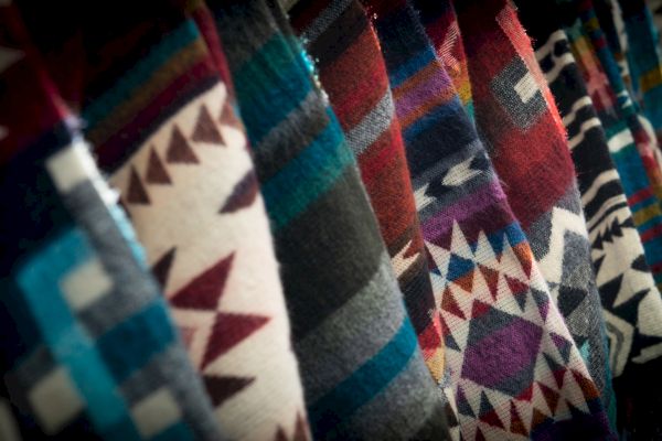 The image shows a row of colorful, patterned fabrics with geometric designs, possibly woven blankets or textiles, displayed vertically.