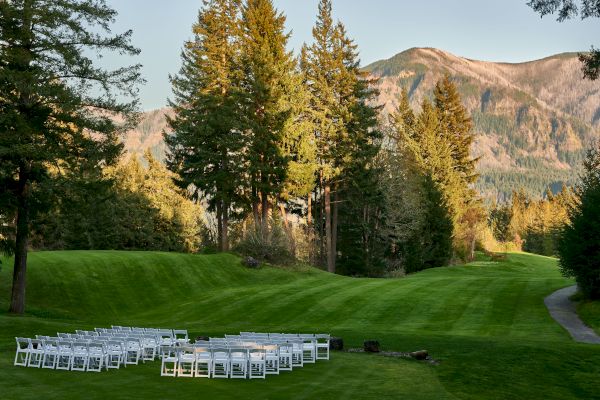 A grassy outdoor setting with rows of white chairs, surrounded by trees and mountains, likely prepared for an event or gathering.