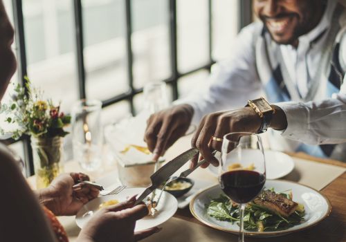 Two people enjoy a meal together at a restaurant, one serving food onto the other's plate, with wine glasses and dishes on the table.