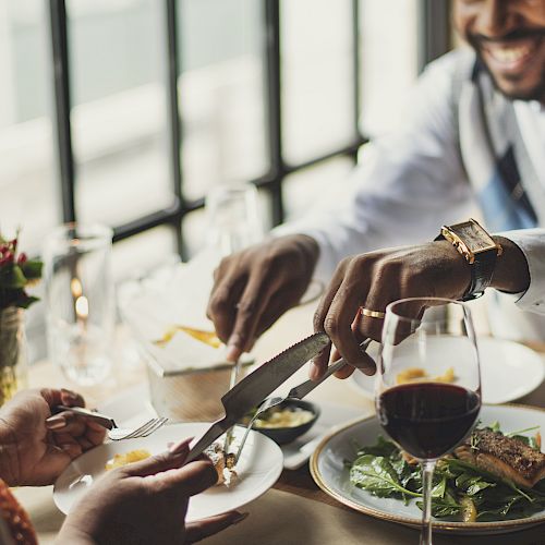 Two people enjoy a meal together at a restaurant, one serving food onto the other's plate, with wine glasses and dishes on the table.