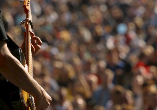 A person is playing a guitar in the foreground, with a blurred crowd in the background, possibly at an outdoor concert or festival.