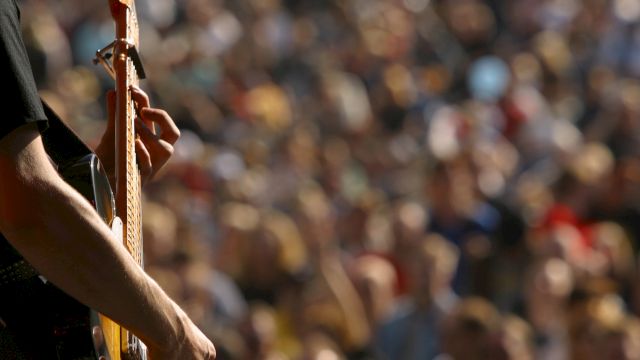 A person is playing a guitar in the foreground, with a blurred crowd in the background, possibly at an outdoor concert or festival.