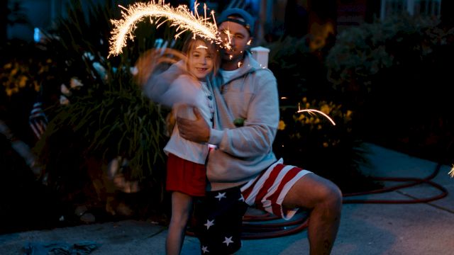 A person kneels, holding a young child with a sparkler making light trails in the air, evoking a joyful and festive atmosphere.