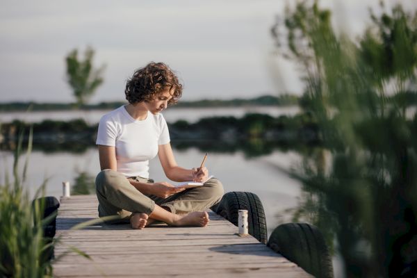 A person is sitting cross-legged on a wooden dock by the water, writing in a notebook, surrounded by nature.
