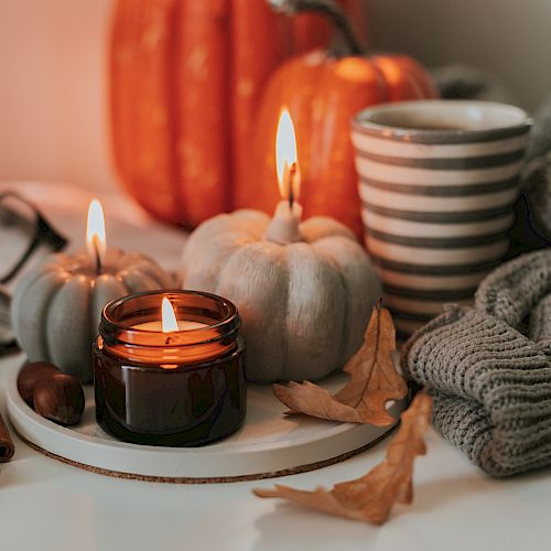Cozy autumn scene with lit candles, pumpkins, a striped mug, knitted blanket, an open book, glasses, and cinnamon sticks on a white surface.