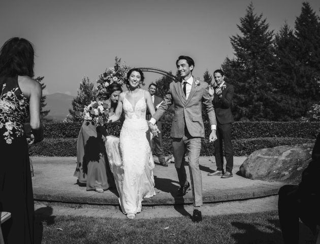 A couple, likely just married, walks down an outdoor aisle, smiling and holding hands, surrounded by guests and greenery.