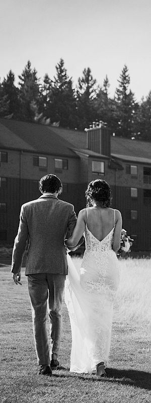 A couple in wedding attire walks toward a building through a grassy field, surrounded by trees, captured in black and white.