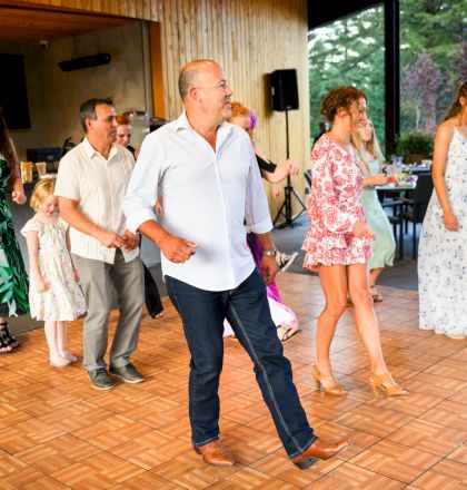 A group of people dancing on a wooden floor at an indoor event; some are dressed casually, others in summer attire.