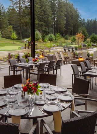 An outdoor dining area with round tables set with plates, glasses, and flowers. Surrounded by lush greenery and trees.