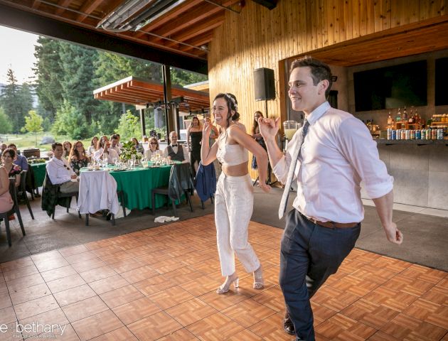 A couple is dancing on a wooden floor at an outdoor event, with guests watching in the background.