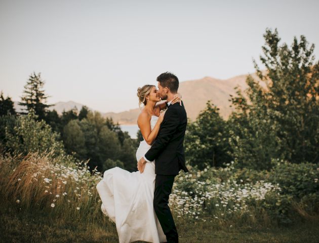 A couple in formal attire embraces in a natural setting with trees and mountains in the background.