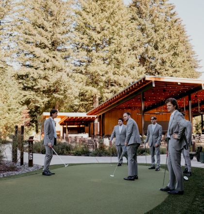 A group of people in suits are playing mini-golf on a green next to a wooden pavilion, surrounded by trees under a clear sky.
