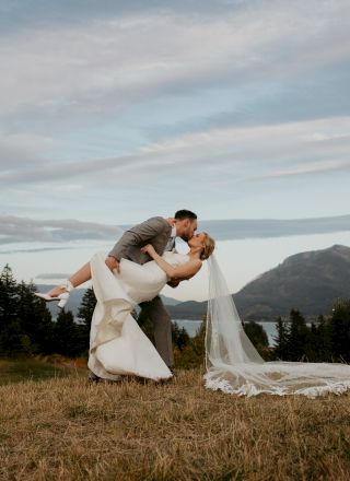 A couple in wedding attire shares a romantic dip kiss in a picturesque outdoor setting with mountains and trees in the background.