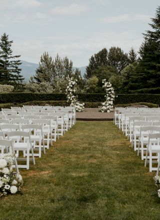An outdoor wedding setup with white chairs and floral arrangements, facing a scenic view of trees and hills in the background.