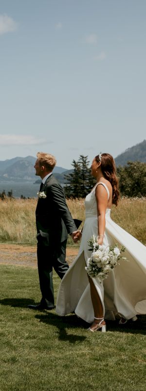 A couple in wedding attire holds hands in a scenic outdoor location with mountains, trees, and a clear sky in the background.