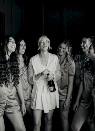 A group of people, dressed in casual and bridal attire, are joyfully gathered indoors with one holding a champagne bottle.